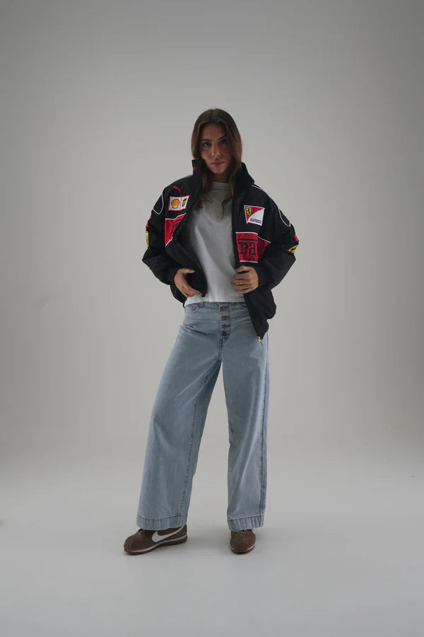 Person wearing a black ferrari jacket with colorful patches, gray shirt, and blue jeans on a plain background