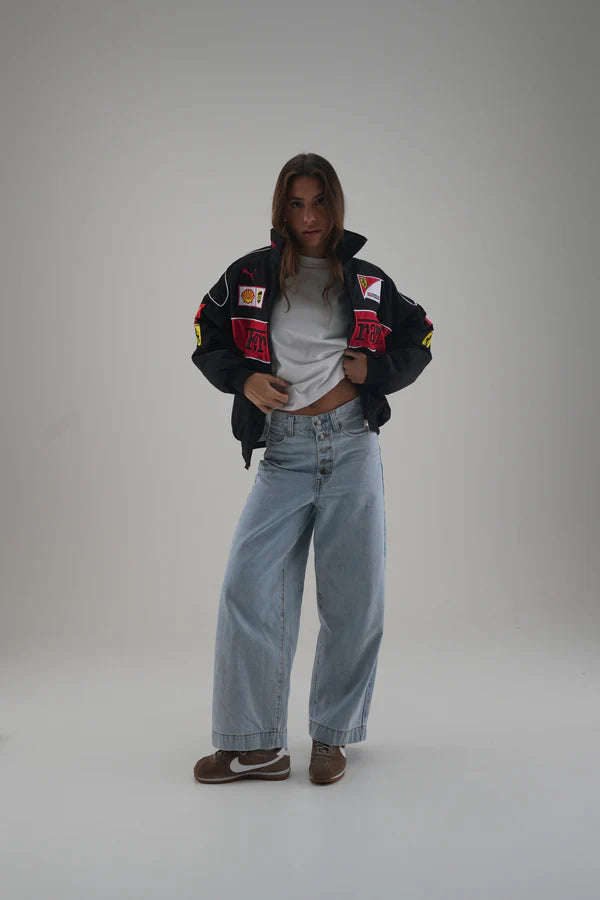Person wearing a black ferrari jacket with colorful patches, light blue jeans, and brown shoes on a gray background