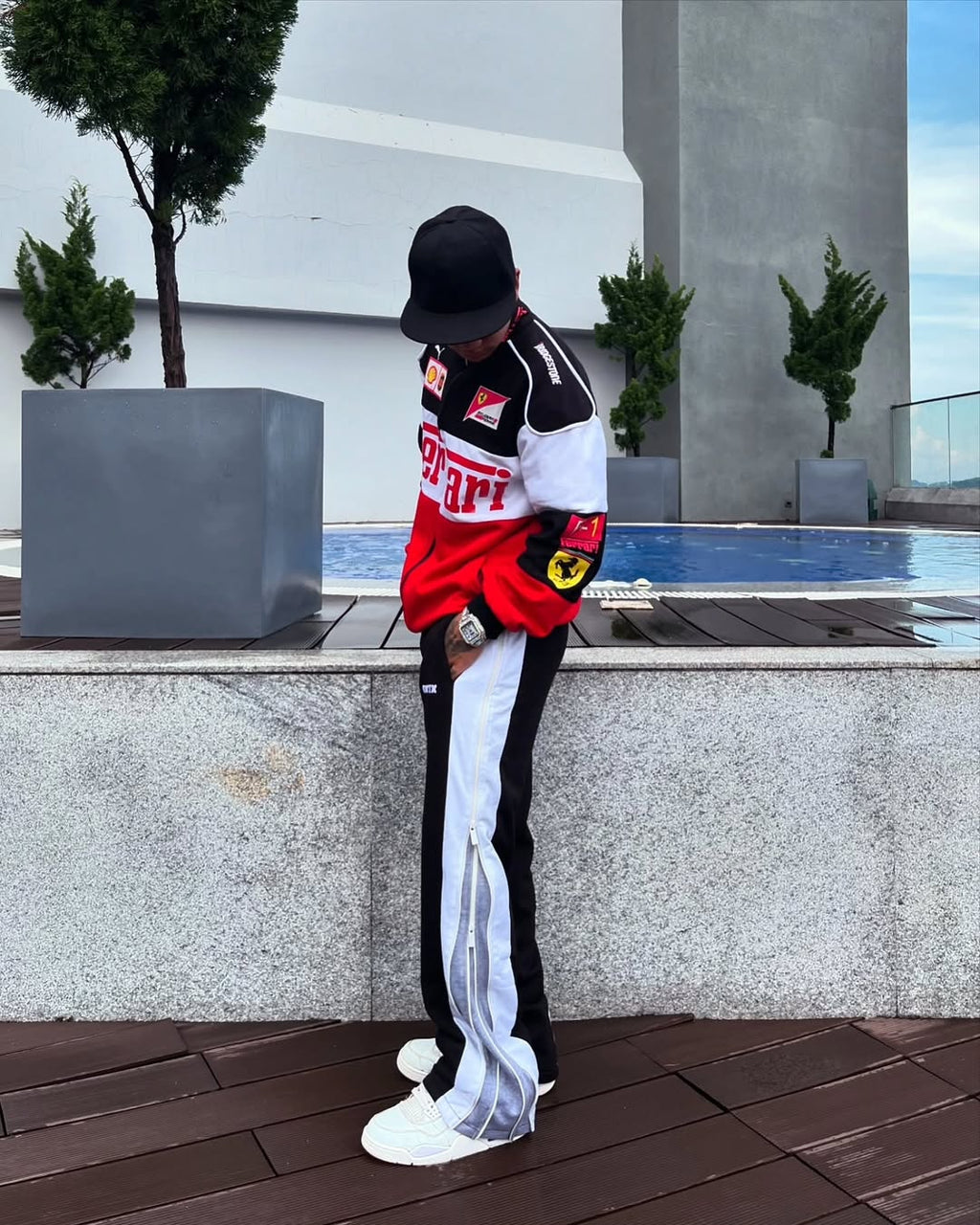 Person wearing a Ferrari tracksuit leaning against a wall with plants and a pool in the background.