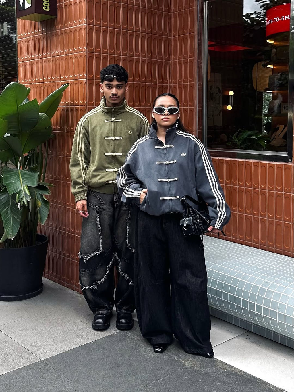 Urban lifestyle shot of the male and female models flexing the Olive Green and Dark Grey CNY bomber jackets. Captures the high fashion 'model off-duty' aesthetic blended with raw Gen Z street culture styling.