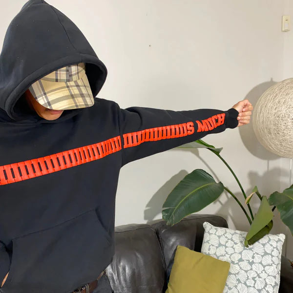 Person wearing a black hoodie with orange text, standing indoors with a plant and sofa in the background.