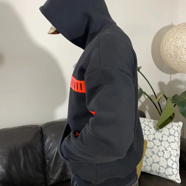 Person wearing a dark hoodie with a red stripe in a room with a couch and lamp.