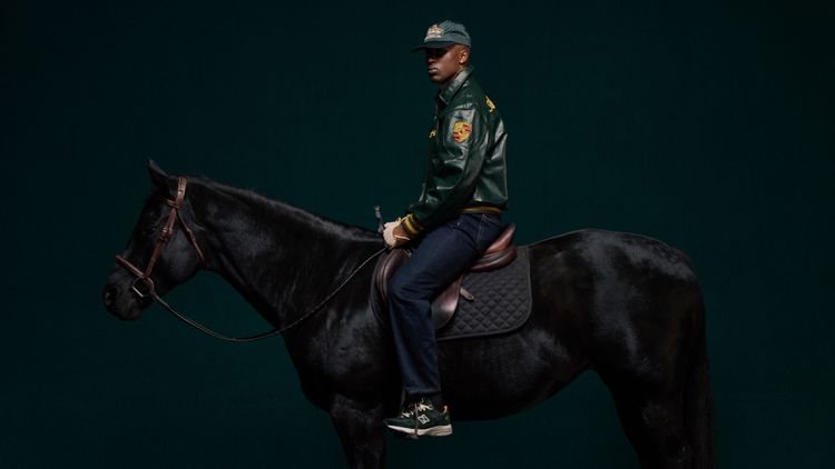 Full body outdoor lifestyle shot of the model wearing the green Porsche 993 faux leather jacket. Showcases the complete Paddock leather jacket streetwear outfit, capturing the incredibly bold unisex streetwear racing aesthetic.