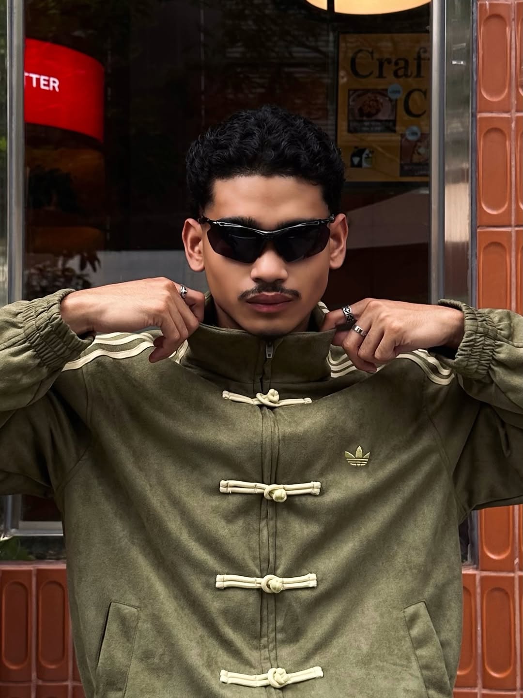 Side profile of the male model demonstrating the masculine box-cut architecture of the Olive Green Custom Knot jacket. Highlights the thick 3-stripe embroidered sleeve detailing synonymous with vintage varsity racing jackets.