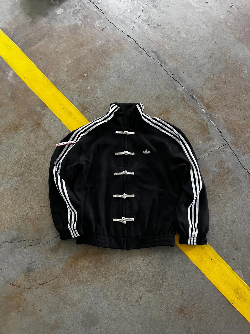 Front view mockup of the Unisex Black CNY Adidas Knot Jacket, displaying the Tang-style design, sleeve stripes, and frog closures