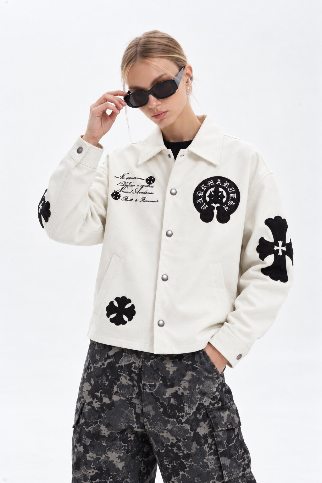 Lifestyle streetwear shot of a girl wearing the cream cross patch leather jacket paired with camouflage cargo pants and dark sunglasses